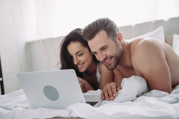 krásná žena a pohledný muž ležící na posteli a používající přenosný počítač  - Fotografie, Obrázek