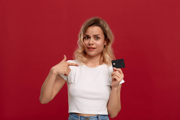 Portrait of a beautiful girl with curly blond hair dressed in a white t-shirt standing on a red background. Model holds bank card and points at it with finger  - Valokuva, kuva
