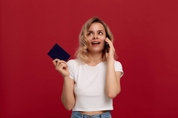 Portret van een meisje met krullend blond haar in een wit t-shirt op een rode achtergrond. Gelukkig model spreekt op de telefoon houder paspoort. - Foto, afbeelding