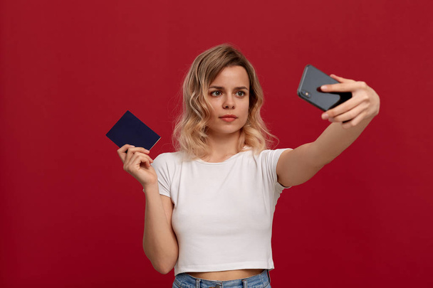 Portrait of a girl with curly blond hair in a white t-shirt on a red background. Model makes selfie with the mobile phone holding passport. - Фото, изображение