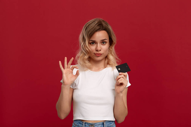 Portret van een meisje met krullend blond haar in een wit t-shirt staande op een rode achtergrond. Model glimlacht op de camera, houdt bankkaart bedekt de helft van een gezicht en toont gebaar van OK-symbool. - Foto, afbeelding