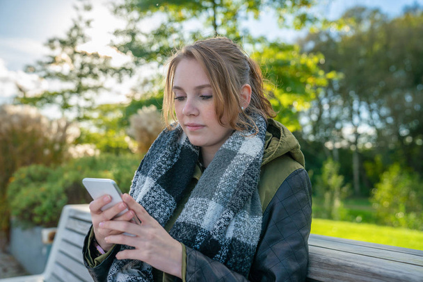 Una joven está sentada en un banco del parque y está escribiendo un mensaje de texto
 - Foto, imagen