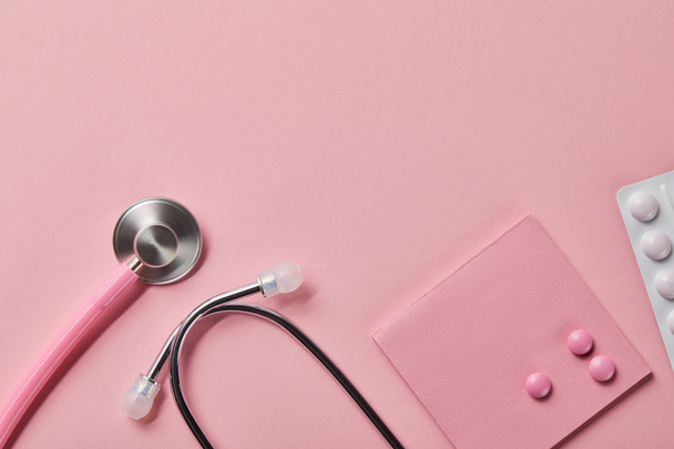 ピンクの聴診器のトップビュー、薄いピンクの背景に紙とブリスターパックの3つの錠剤 - 写真・画像