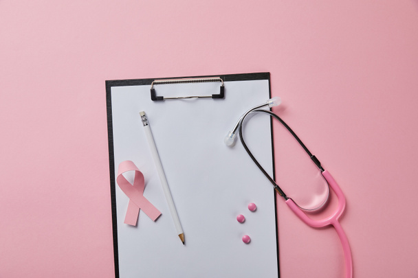 pencil, pink pills and breast cancer ribbon on folder with empty paper near stethoscope on pink background - Photo, Image