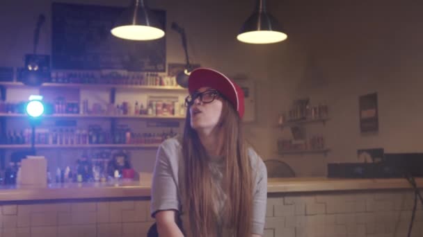 Uma jovem bonita de boné vermelho fuma um cigarro eletrônico na loja de vape. Fecha a porta. Movimento lento
. - Filmagem, Vídeo