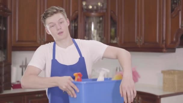Portrait young handsome smiling man taking the basket with detergents and wiping his forehead with his hand. Cleaning day. Housekeeping household housework and cleaning service concept - Кадри, відео