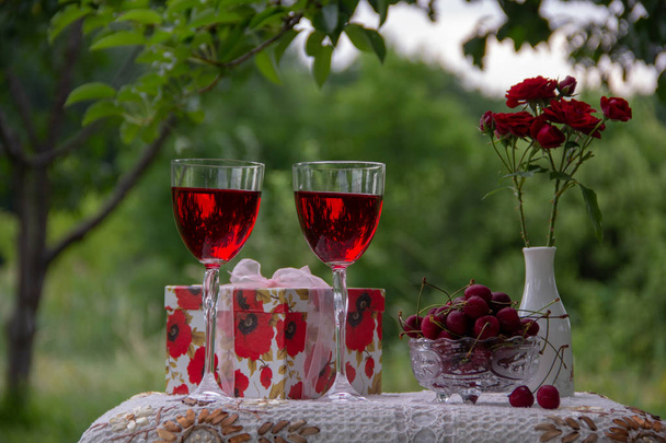 赤い飲み物を持つ2つのグラス、熟したサクランボのクリスタルの花瓶、赤いバラの花瓶、緑の庭に美しいテーブルクロスで飾られたテーブルの上にギフトスタンド付きのボックス. - 写真・画像