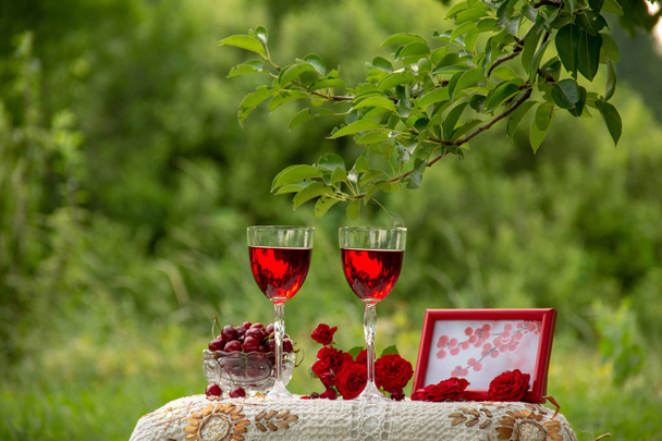 赤い飲み物を持つ2つのグラス、熟したサクランボとクリスタルの花瓶、赤いバラの花束、写真付きのフォトフレーム、自然の緑の庭で美しいテーブルクロスで飾られたテーブルの上に立っています. - 写真・画像