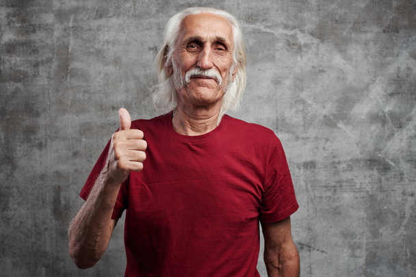 Modern gray-haired caucasian old man with a mustache in red t-shirt shows thumb up and smiles, joyful happy face against gray wall - Photo, image