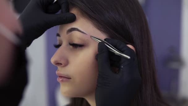 Permanent makeup. Beautiful young woman gets eyebrow correction procedure. Beautician hands in black gloves are tweezing clients eyebrows in beauty saloon - Záběry, video