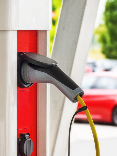 電気自動車用の充電ケーブルを赤白の充電ステーションに差し込み、車が背景に見える. - 写真・画像