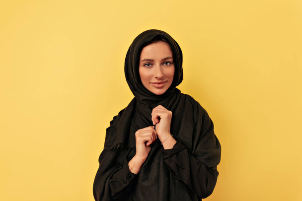 Studiový portrét půvabné arabské ženy stojící se založenýma rukama a s pohledem na fotoaparát izolovaný přes žluté pozadí - Fotografie, Obrázek