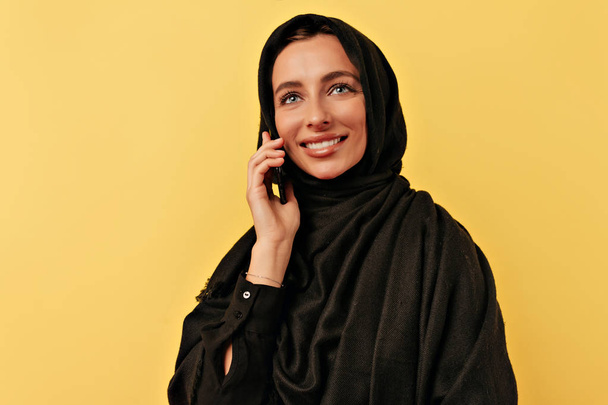 Close up portrait of arabian woman talking on smartphone with charming smile wearing burqa over yellow background - Photo, image
