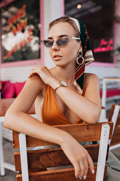 Retrato exterior de mujer atractiva y elegante con gafas y chal en la cabeza sentado en la terraza de verano
  - Foto, imagen