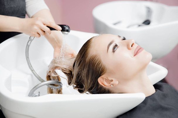 Concept beauty salon. Hairdresser washes hair of beautiful blonde girl under tap in wash - Fotoğraf, Görsel