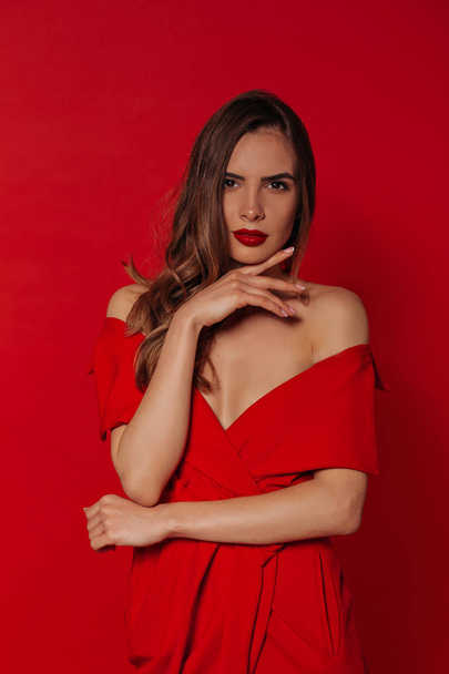 写真撮影中に赤い背景の上にスタイリッシュな赤いドレスを着て赤い唇を持つ自信のある現代の美しい女性 - 写真・画像