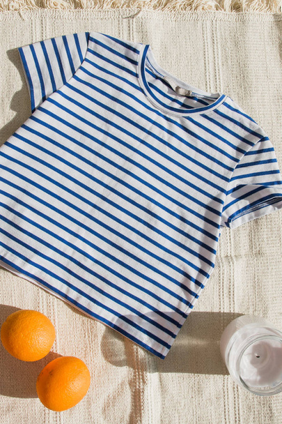 La parte superior recortada de moda con rayas azules se encuentra en la alfombra ligera. Camisa junto a una naranja y una vela. Chaleco con estilo se encuentra sobre un fondo claro
 - Foto, Imagen