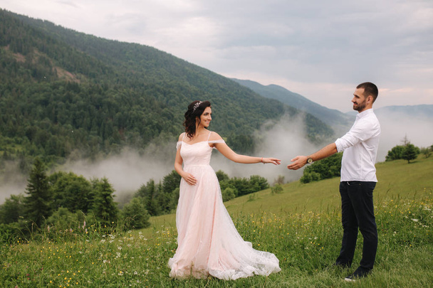Hermosa joven pareja de boda de pie en la pendiente verde, colina. Novio y novia en las montañas Cárpatos
 - Foto, Imagen