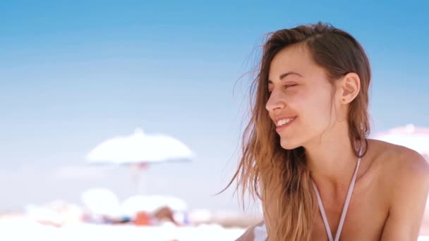 美しい日焼けした笑顔の女性が白い砂浜にビーチに横たわっている、リラックスして小さなヘッドフォンで携帯電話から音楽を聴きます。ビキニとホワイトキャップ enjoing とトプリーのスリムセクシーな女性 - 映像、動画
