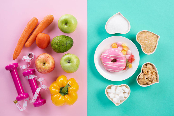 Healthy lifestyle, food and sport concept. Top view of healthy versus unhealthy. Donut and various types of sugar VS fruit vegetables, measuring tape and dumbbell on pink blue pastel background. - Photo, Image