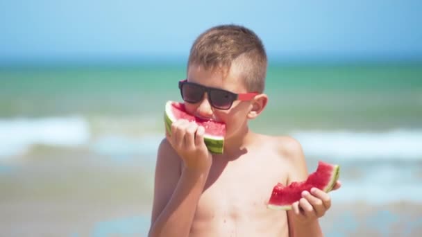 Το αγόρι με τα μαύρα γυαλιά τρώει καρπούζι στη θάλασσα. - Πλάνα, βίντεο