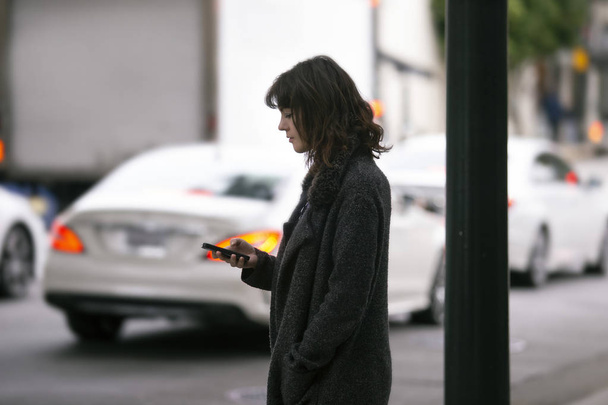 Female pedestrian waiting on a sidewalk for a rideshare.  She is sharing her gps location via cellphone app so the driver can pick her up in the city.  Cars are blurred to obscure make model and license plates.  - Photo, image