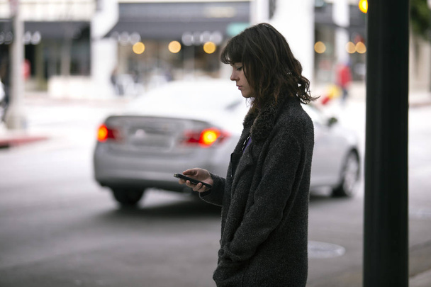 Female pedestrian waiting on a sidewalk for a rideshare.  She is sharing her gps location via cellphone app so the driver can pick her up in the city.  Cars are blurred to obscure make model and license plates.  - Фото, изображение