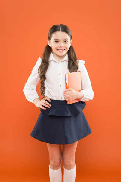 Schoolgirl studying textbook. Interesting book for children. Homeschooling concept. small girl in school uniform. schoolgirl writing notes on orange background. using textbook at school lesson - Photo, image