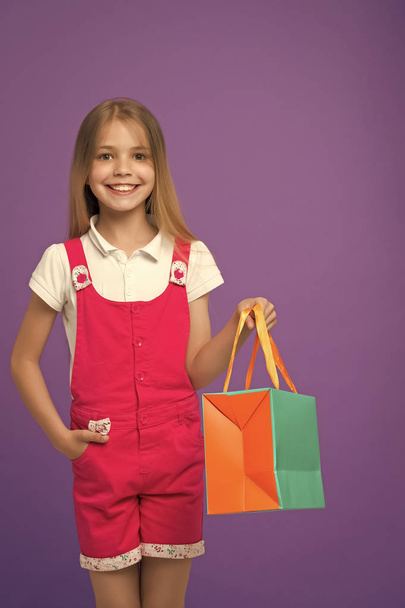Happy girl with shopping bag on violet background. Little shopaholic smile with paper bag. Child shopper in fashion jumpsuit. Holidays preparation and celebration. Childhood. Shopping and sale - Photo, Image