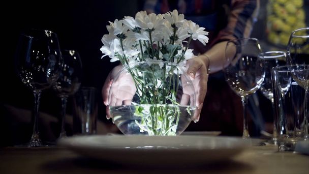 Woman puts vase of flowers on table. Action. Close-up of woman puts vase of white flowers on serving table with mysterious lights on dark background - Photo, Image