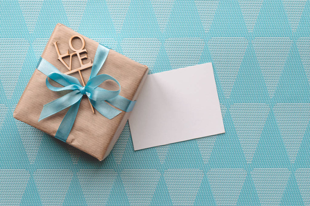 Caja de regalo envuelta en papel marrón con cinta azul claro sobre fondo azul con tarjeta de felicitación. Burlarse. Espacio libre
 - Foto, imagen