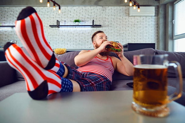 Thick funny man eating a burger sitting on the couch. - Foto, Imagem