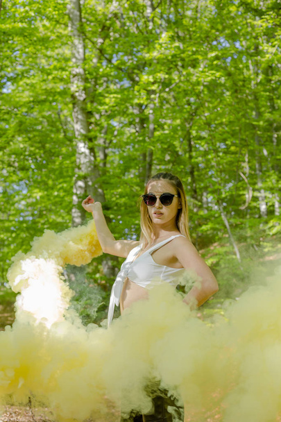 junge Frau in Sonnenbrille und Sommerkleidung mit Rauchbombe im sonnigen Wald  - Foto, Bild