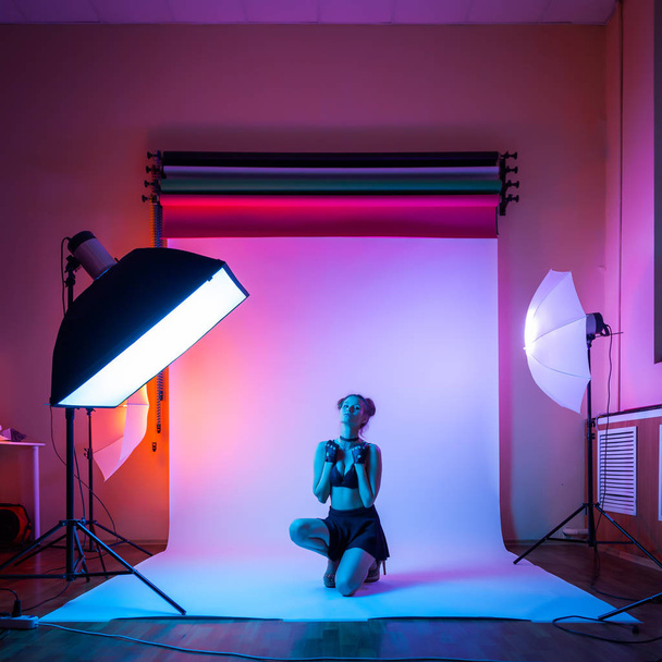 Backstage: model woman with in black underwear, pantyhose, skirt posing in attractive position in colorful bright neon blue and pink lights in studio. Fashion concept - 写真・画像
