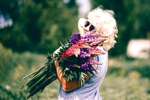 Blonde mature femme robuste tenant un bouquet de lupin sur fond de nature. Le concept de beauté et de corps positif. Lumière du coucher du soleil
 - Photo, image