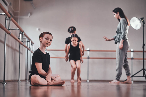 Girl is sitting on the floor while her other classmates doing exercises with trainer. - Фото, изображение