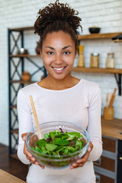 Kaunis mulatto nainen kotona osoittaa terveellistä ruokaa, pitää käsissään salaatti - Kuva
 - Valokuva, kuva