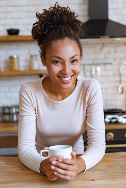 Ładna dziewczyna mulat siedzi przy stole z filiżanką porannej herbaty lub kawy i uśmiechnięty patrząc na aparat fotograficzny - Zdjęcie, obraz
