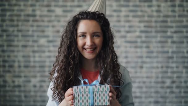 Portrait of cute girl in party hat offering gift box smiling looking at camera - Πλάνα, βίντεο