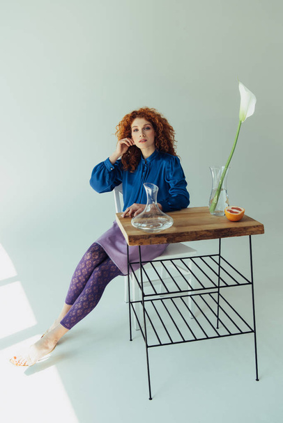beautiful fashionable redhead girl posing at table with calla flower and vases on grey - 写真・画像