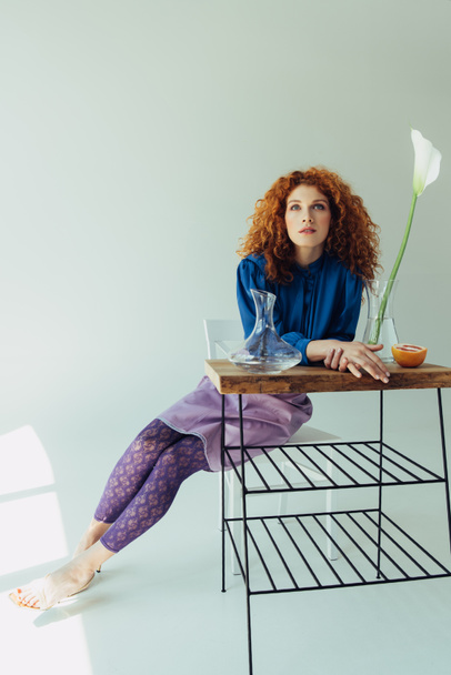 attractive fashionable redhead girl posing at table with calla flower and vases on grey - Fotografie, Obrázek