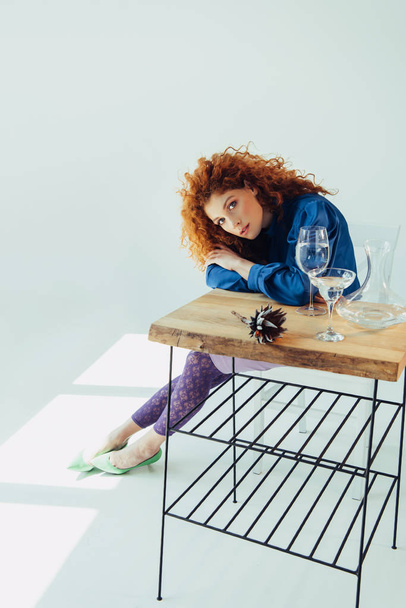 redhead girl looking at camera and posing at table with calla flower and glasses on grey - Foto, afbeelding