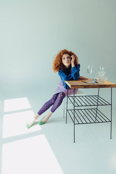 beautiful redhead girl looking at camera and posing at table with calla flower and glasses on grey - Photo, Image