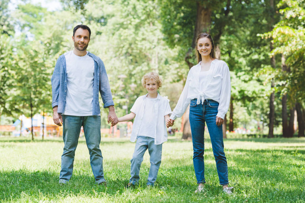 happy family looking at camera and holding hands in park - Photo, Image