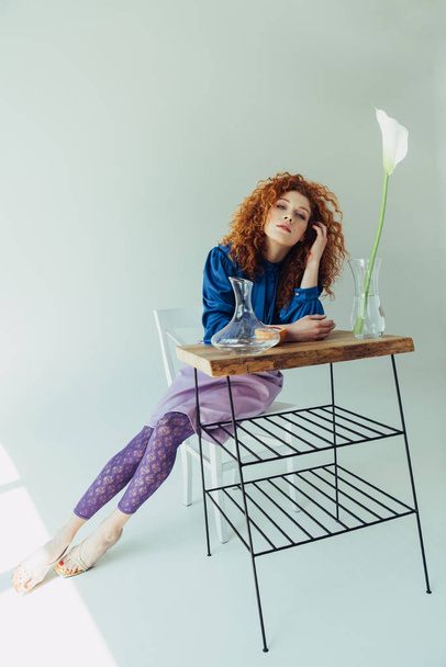 beautiful trendy redhead girl posing at table with calla flower and vases on grey - Photo, Image