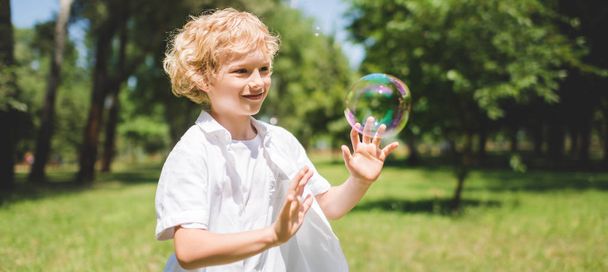 panoramic shot of cute boy Gesturing near soap bubbles in park - Photo, Image