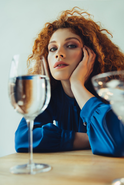 красивая стильная рыжая женщина, подпирающая голову и позирующая возле стаканов, изолированных на сером
 - Фото, изображение
