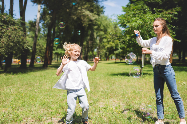krásná mamka fouká mýdlové bubliny pro rozkošného syna v parku - Fotografie, Obrázek