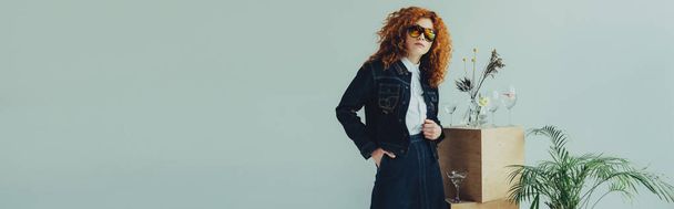 panoramic shot of stylish redhead girl posing near wooden boxes, glasses and plants isolated on grey - 写真・画像