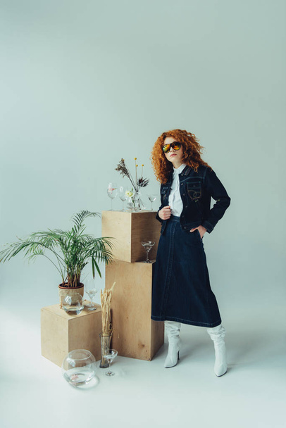 stylish redhead girl posing near wooden boxes, glasses and plants on grey - Photo, image